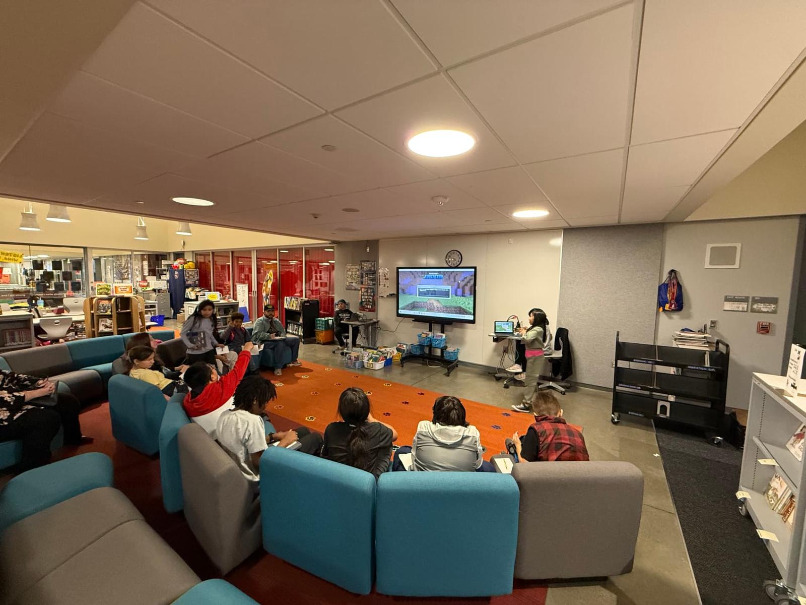 The same kind of room turned into a BlackMuse session: students on soft seating watching a classmate present a game build on the big screen