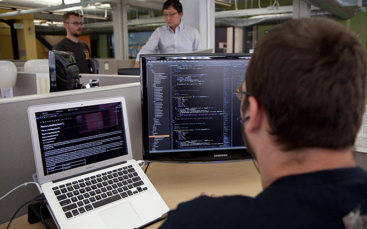 Software developer reviewing code across a laptop and dual monitors at a shared desk