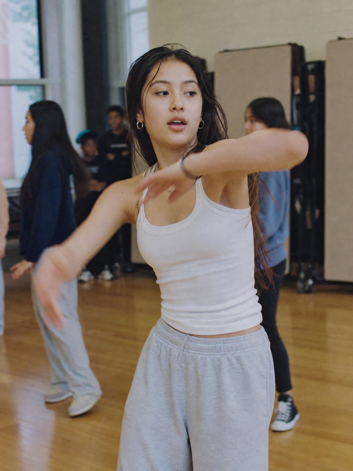 A student mid dance move in a school multipurpose room, classmates practicing behind her