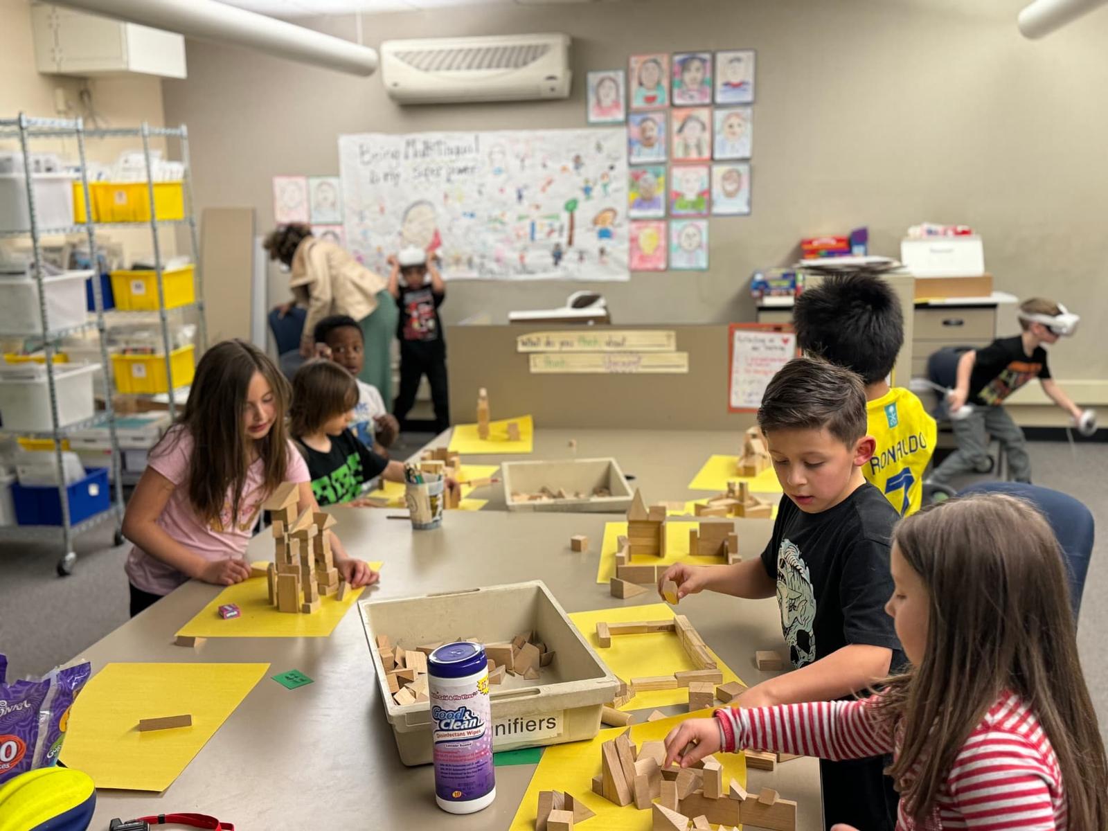 Students build structures with wooden blocks while a classmate explores in a VR headset in the background